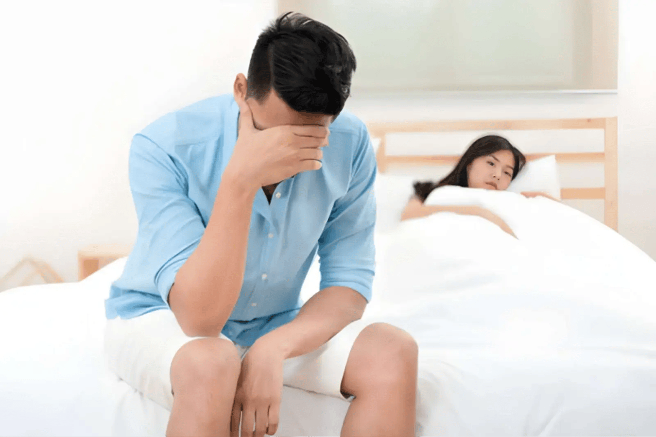 Man sitting on the edge of a bed with his face in his hand while his partner lies awake behind him.